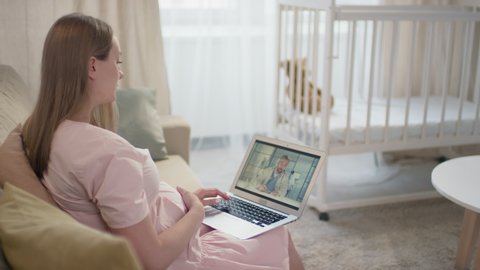 Medium slow-motion side-view footage of young pregnant woman talking with gynecologist by video chat on laptop stroking belly sitting in living room