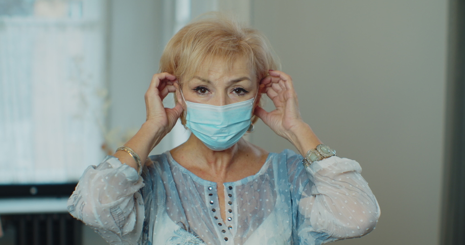 Serious older woman putting surgical mask that protects from epidemic outbreak. Senior woman looking to camera portrait. Social distancing during Covid 19 Coronavirus quarantine lockdown, slow motion. | Shutterstock HD Video #1064741641