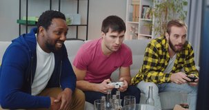 Multi-ethnic male best friends sitting on couch in room and playing video games Caucasian guys spending time playing on console soccer game. African American happy man eating and drinking with company