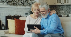 Happy senior old couple laughing talking relaxing on sofa using digital tablet, cheerful mature aged family spending time with gadget having fun.