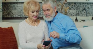 Old elderly adult family couple sitting at sofa with smartphone holding device enjoy internet funny video, search goods on e-commerce, discuss social media news, using modern technology at home