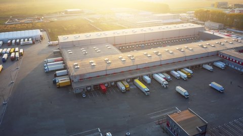Logistics park with a warehouse and loading hub. Semi-trailer trucks with cargo trailers stands at the ramps for load and unload goods at sunset. Aerial hyper lapse.