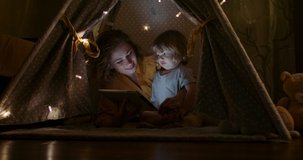 Positive caucasian woman and her son are watching cartoons on a tablet together, happily sniling. Family having fun in cozy tent in evening 4k footage