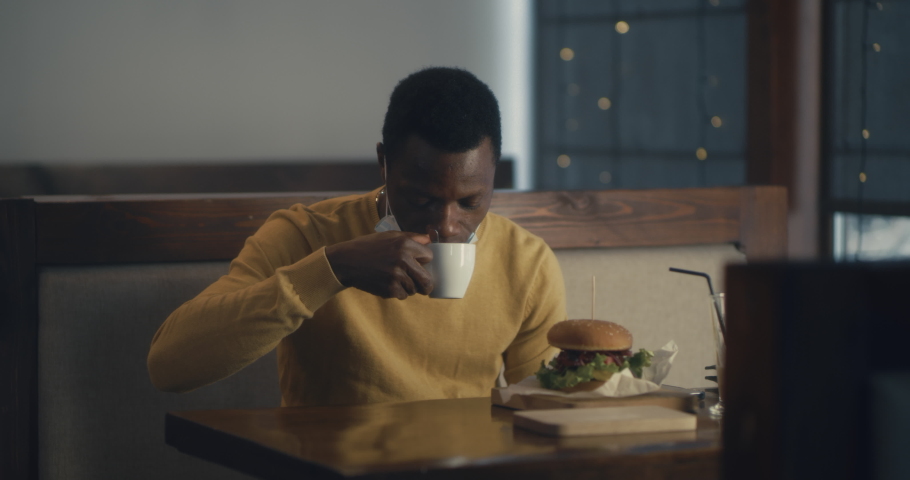 Young African American guy with medical mask having burger and coffee and paying with RFID microchip for meal Royalty-Free Stock Footage #1064750626