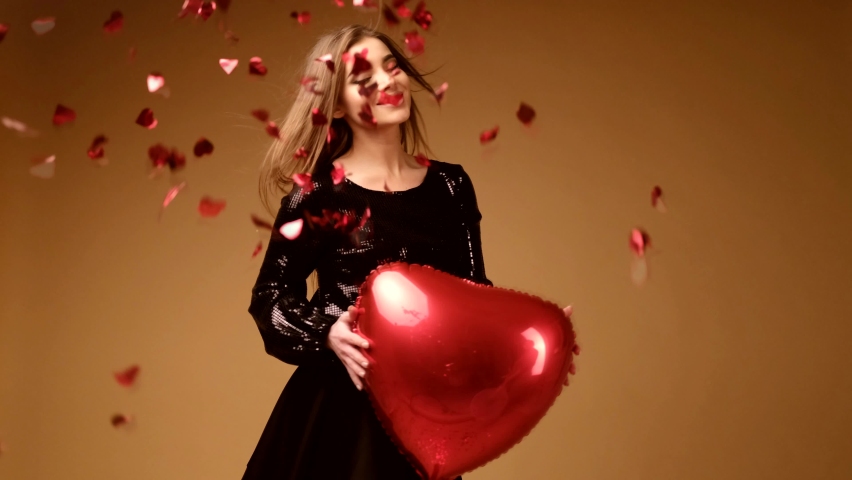 A girl in a black dress with a big red heart in her hands rejoices against the background of falling confetti.