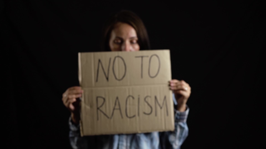 A young beautiful caucasian girl in blue plaid shirt stands against black background holds, his hands piece of brown cardboard with inscription NO TO RACISM. Concept of inequality, social problem