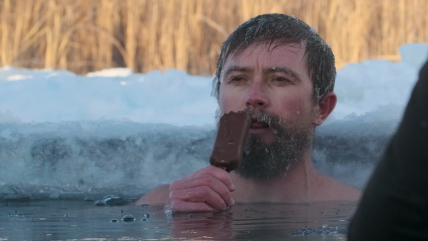 Cryotherapy. Young caucasian man with icy beard calmly eats an ice cream being emerged in the cold icy water
