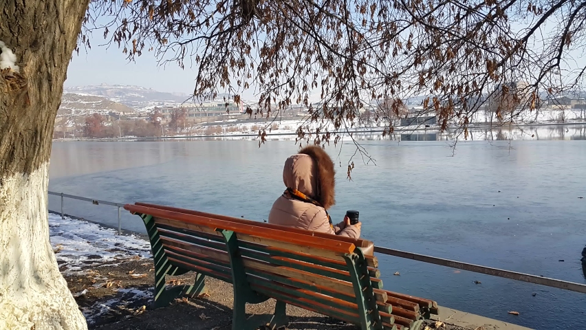 A girl in a winter coat was sitting on a bench near an artificial lake, looking at the frozen lake and drinking hot tea from a thermos.