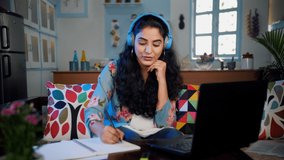 Indian female talking on a video call on the laptop and making her notes. Medium shot of a businesswoman wearing blue headphone sitting on a sofa while talking to her client on the internet