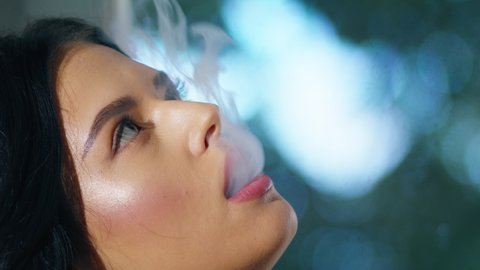 Beautiful young brunette girl sitting and vaping.  Woman puffs on a vape pen device. Blowing smoke out of your mouth deliciously. Close-up cinematic smoking.