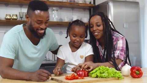 Happy african family in kitchen having fun and cooking together. Portrait of afro-american mother and father teaching little daughter cutting vegetables making dinner together
