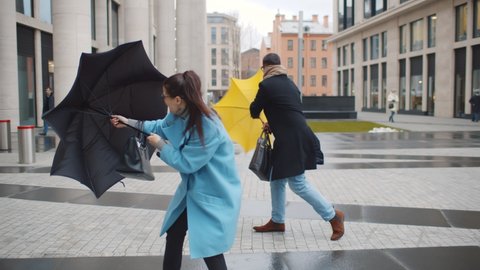 Businessman and businesswoman in coat shelter umbrella from the rain and strong wind outside in bad weather. People on street trying to open umbrella sheltering from stormy wind