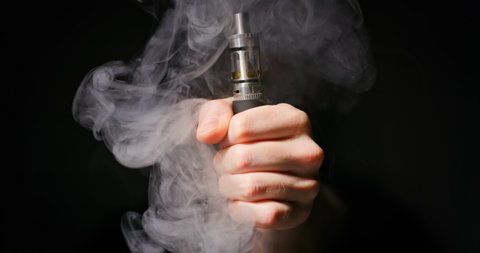 Electronic cigarette grabbed by male hand against dark background and vapor