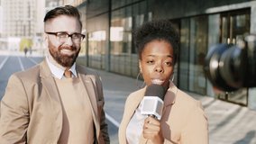 Handsome Caucasian businessman standing outdoors in downtown, smiling and speaking into microphone on camera while giving interview to Afro-American female journalist