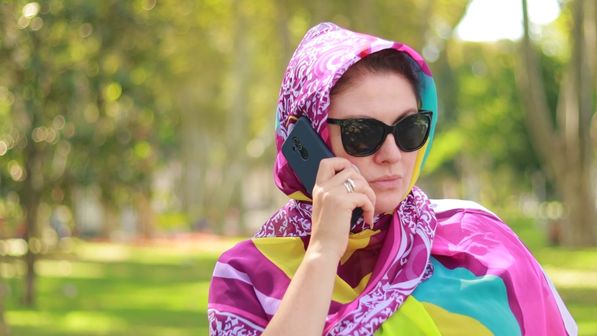 A young woman in a headscarf is talking and taking a photo with a smartphone in her hands. Summer sunny day in the park.