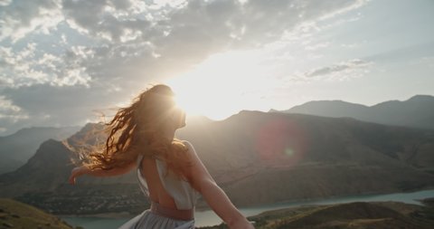 Woman in white dress standing on top of a mountain with raised hands while wind is blowing her dress and red hair - freedom, nature concept 4k footage