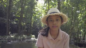 Portrait cute girl wearing straw hat is smiling and looking at video camera while staying at tropical forest during summer vacation. Thailand.
