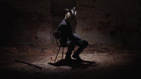 Possessed maniac tied to a chair. Broken light over a tortured person in animal mask. Eerie, frightening scene, scary basement.