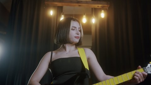 Young beautiful woman with short hair in a black dress plays the electric guitar in a modern venue