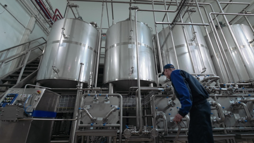 Male factory worker at modern drink production plant controls the advanced equipment. Stainless huge containers at modern beverage production factory. Soft drink machinery system line. Slow motion Royalty-Free Stock Footage #1064811217