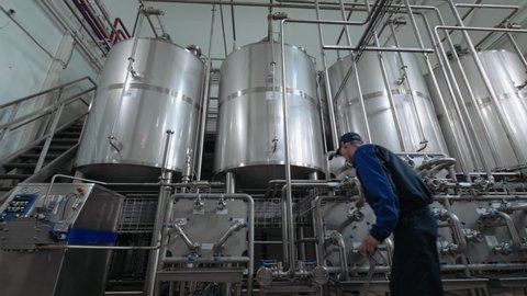 Male factory worker at modern drink production plant controls the advanced equipment. Stainless huge containers at modern beverage production factory. Soft drink machinery system line. Slow motion