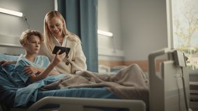 Hospital Ward: Handsome Young Boy Resting in Bed,  His Beautiful Mother Visiting Uses Smartphone for Video Call Conference Talk with Family and Friends. Happy Patient Recovering after Sickness