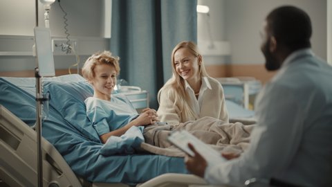 Hospital Ward: Handsome Young Boy Resting in Bed with Caring Mother Visits to Support Him, Friendly Doctor Talks, Gives Advice. Happy Smiling Patient Recovering after Sickness or Successful Surgery