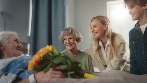 Hospital Ward: Grandfather Resting in Bed, His Caring Beautiful Grandmother Sitting Beside, Happy Grandson and Daughter Visit, Hug Lovingly, Give Flowers. Family Support Old Man Recovering