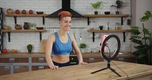 Smiling woman blogger with stylish haircut and bare tummy shoots video for blog with phone and light ring in spacious kitchen