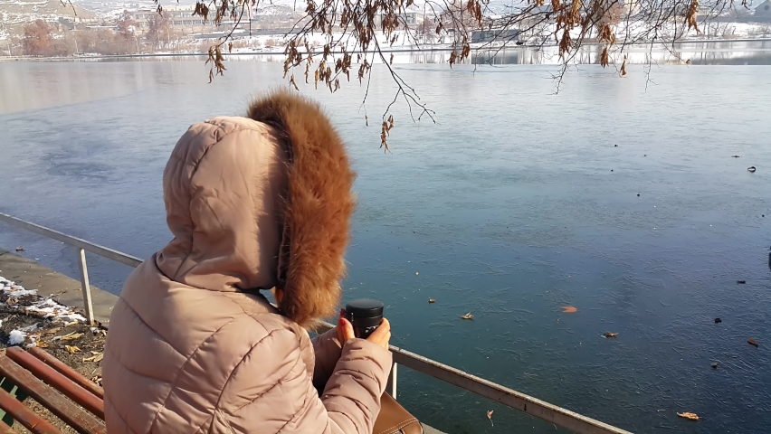 A girl in a winter coat was sitting on a bench near an artificial lake, looking at the frozen lake and drinking hot tea from a thermos.