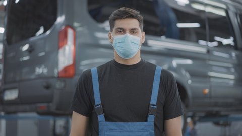 Confident man in coronavirus face mask walking in slowmo to camera taking off working gloves. Portrait of confident handsome Caucasian auto mechanic working in repair shop on Covid-19 pandemic.