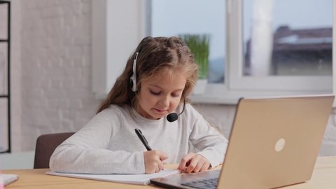Schoolgirl in headset has online lesson, video call zoom with teacher, answering questions,check of knowledge.Cute primary school girl studying at home using laptop. Distance learning. Home education.