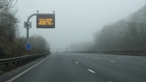 Driving highway in fog; there is a message on roadside board for drivers saying: Stay home, essential travel only. Due to coronavirus pandemic in the UK travelling has restrictions.