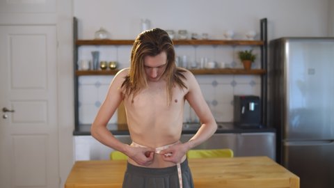 Young thin shirtless man measuring waist with tape at home. Portrait of handsome guy on diet using measuring tape standing in modern kitchen