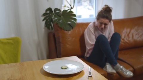 Focus on plate with cucumber slice at table and crying depressed woman on background. Frustrated and sad young female sitting on couch and crying suffering from eating disorder
