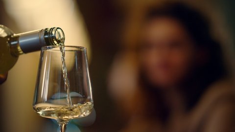 Closeup unknown waiter hand pouring wine into glass indoors. Young woman hand taking wineglass with drink in restaurant interior. Unrecognizable bartender hand holding bottle of white wine in bar.