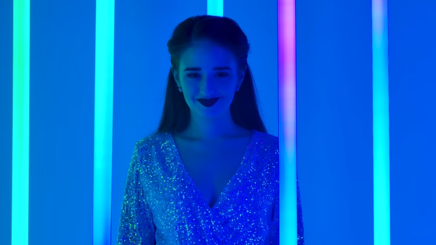 Portrait of a charming young woman laughing happily in the studio surrounded by colorful neon tubes. The girl puts her hands together, expressing her delight. Close up. Slow motion.