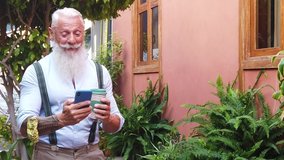 Senior hipster man holding coffee and having fun with a modern phone on a europen city street, close up view, happiness, technology and elderly lifestyle people concept