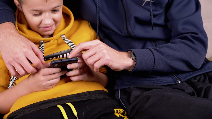 Happy son with dad laughing looking at screen smartphone, father teaching boy using apps sitting on sofa. Hands holding smartphone, dad talking to son, pointing finger at screen. Happy family. 4K. | Shutterstock HD Video #1064872678
