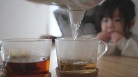 Pour boiling water from an electric kettle pot into glass cup containing an Earl Gray tea bag. Asian little toddler girl watching while eating snack next to her mother in the background. Wooden table - Powered by Shutterstock - Get 15% off with code: PIKWIZARD15