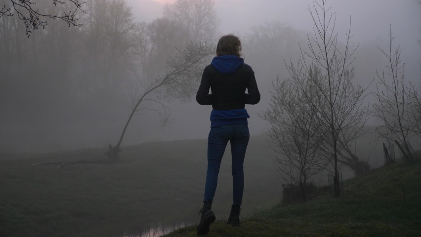 Young lost woman looks around in mysterious forest. Female surrounded by fog and dark evening sky. Grainy view.