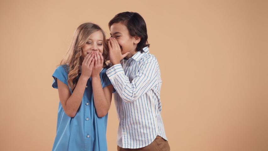 A cute boy in a striped shirt whispers a joke or gossip to a cute girl in the ear with curly hair in a blue dress and they laugh together isolated on a peach tender background.Valentine's Day. | Shutterstock HD Video #1064878081