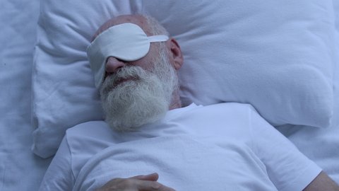 Worried greyhaired male wearing eye mask, trying to sleep at night, insomnia