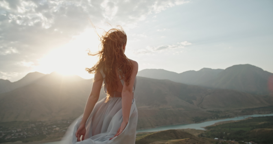 Woman in white dress standing on top of a mountain with raised hands while wind is blowing her dress and red hair - freedom, nature concept 4k footage | Shutterstock HD Video #1064893954
