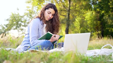 Young adult girl student studying in nature in the park on the lawn using a laptop on a sunny summer day. Pretty and attractive female freelancer working at a distance online Distances. Relax and rest