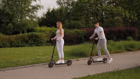 Young beautiful guy and girl in white clothes ride electric scooters in the Park at sunset.