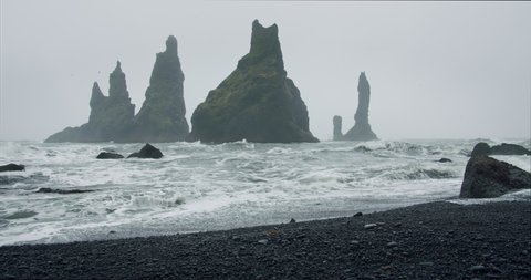 Sharp cliffs on black sand beach of Reynisfjara with waves hitting the shore on foggy rainy stormy day. Vik, Iceland.