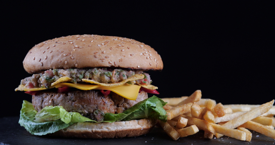 Rotating shot of appetizing double Burger with meat beef salad tomatoes cheese and onions stands on black table hamburger with fries and chips against dark background close-up, American tradition food
