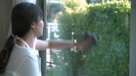 Happy young Asian housewife - maid using a glass cleaning tools wipe on the glass window surface with liquid cleaning solution or detergent. Housekeeper cleaning a dirty glass window close up.