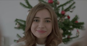 Camera view of young caucasian female using smart phone video call talking with friends, Christmas tree decorated with ornament in living room at home. Social distancing, New Year holiday festival.